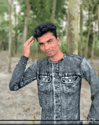 a man in a denim jacket stands in the woods with his hand on his forehead