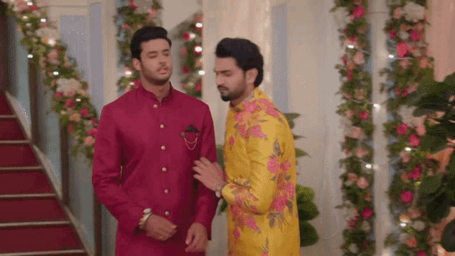 two men in traditional clothes are standing next to each other in a room with flowers .