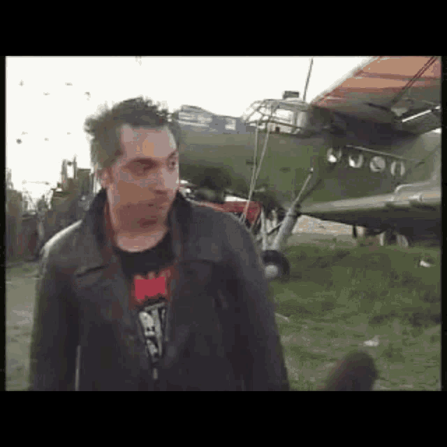 a man wearing a black jacket with the word ny on it stands in front of an airplane
