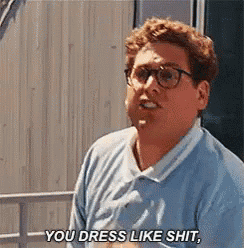 a man wearing glasses and a blue shirt is standing in front of a building and saying `` you dress like shit , '' .