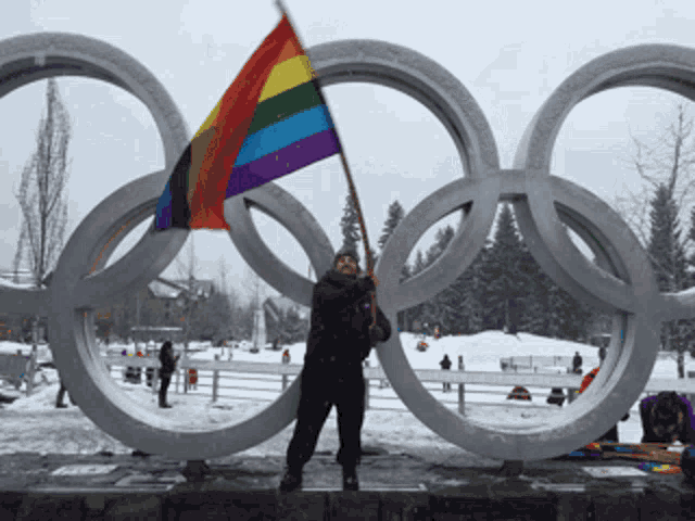 a man holds a rainbow flag in front of the olympic rings in the snow