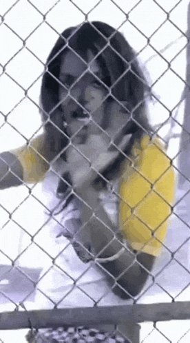 a woman in a yellow shirt is behind a chain link fence making a funny face