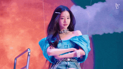 a woman in a blue off the shoulder top and denim shorts is standing on a stage with her arms crossed .
