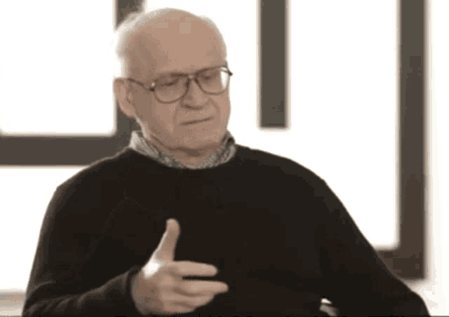 an older man wearing glasses and a black sweater giving a thumbs up