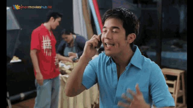 a man in a blue shirt is talking on a cell phone in front of a sign that says pinoymovieshub.net