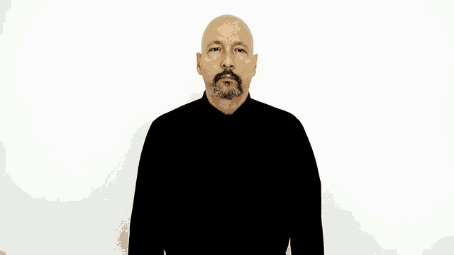 a bald man with a beard is wearing a black turtleneck and making a hand gesture on his forehead .