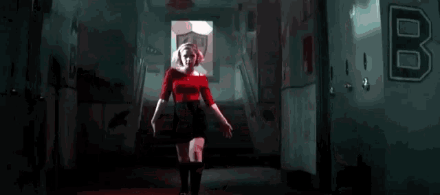 a woman in a red top and black skirt is standing in a dark hallway holding a knife .