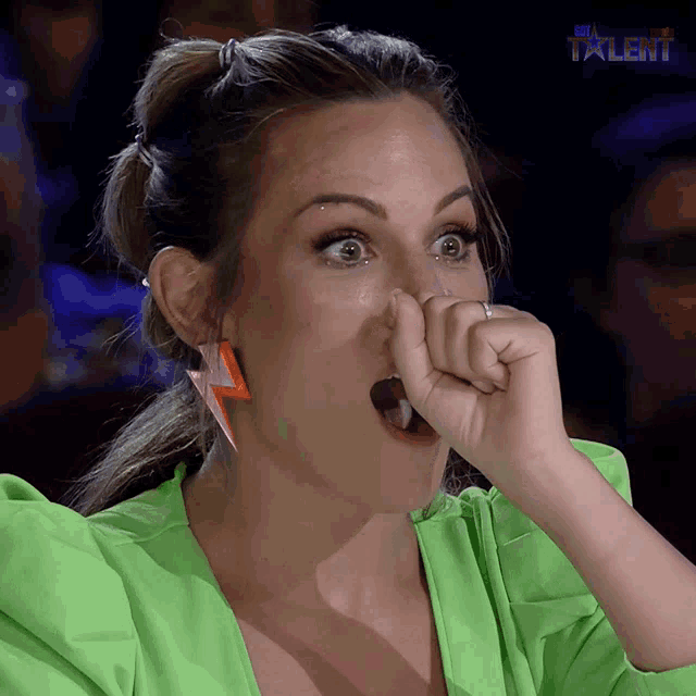a woman in a green jacket is covering her nose with her hand while watching a talent show