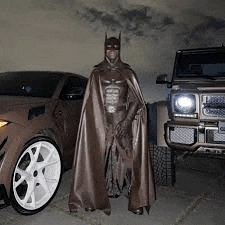 a man in a batman costume is standing in front of a car and a jeep .