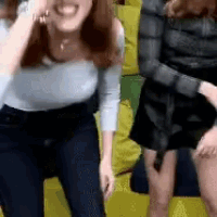 two women are standing next to each other on a yellow couch and laughing .