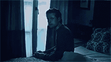 a man is sitting on a bed in front of a window looking out .