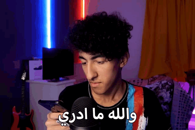 a young man with curly hair is holding a cell phone in front of a microphone with arabic writing on his shirt