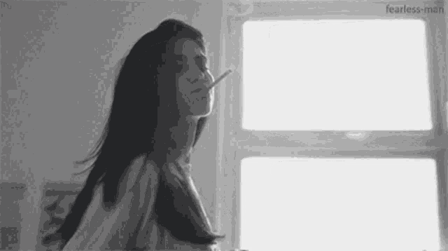 a black and white photo of a woman smoking a cigarette in front of a window .