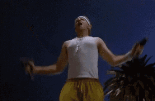 a man in a white tank top and yellow shorts is holding a flamethrower in his hand .