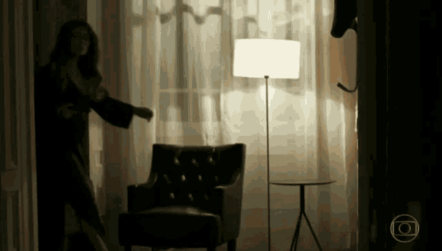 a woman is standing in a dark room with a lamp and a chair