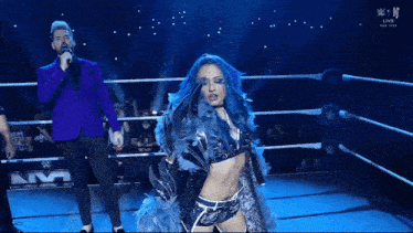 a woman with blue hair stands in a wrestling ring while a man holds a microphone