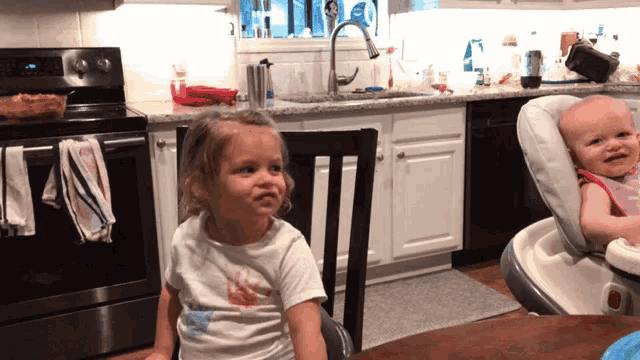 a little girl sits at a table while a baby sits in a high chair in a kitchen