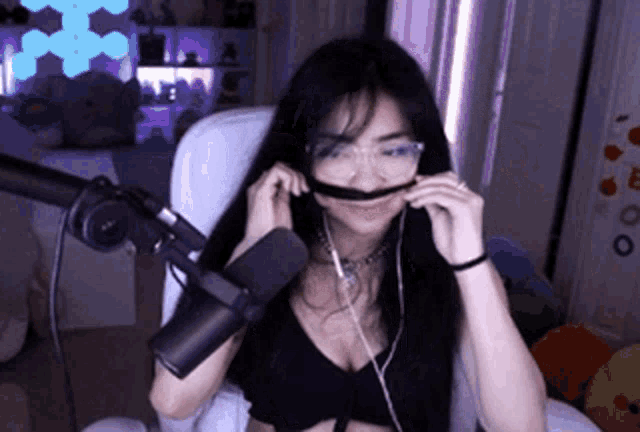 a woman wearing glasses and headphones is sitting in front of a microphone and making a mustache .
