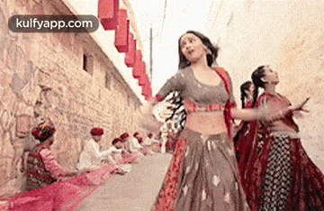 a group of women are dancing in a street while men sit on the ground .