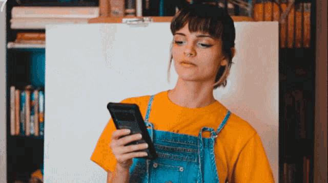 a woman in overalls and a yellow shirt looks at her phone
