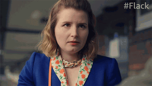 a woman wearing a blue jacket and a floral shirt is looking at the camera with the hashtag #flack behind her