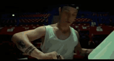 a man in a white tank top is sitting in a dark room with a tattoo on his arm .