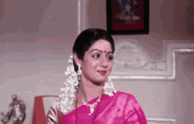a woman in a pink saree with white flowers in her hair looks at the camera .