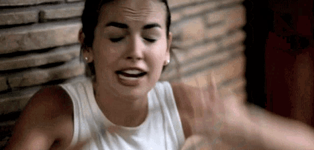 a woman in a white tank top is sitting in front of a brick wall and making a face .
