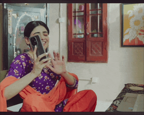 a woman in a purple and orange floral dress is sitting on a bed holding a cell phone