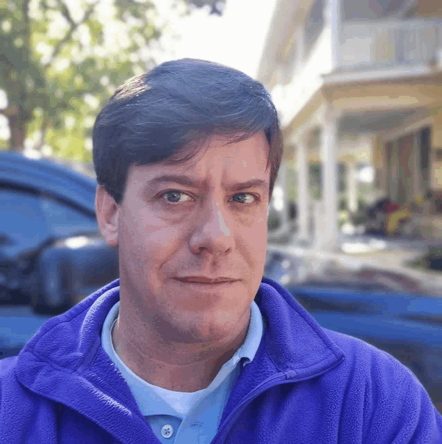 a man in a purple jacket looks at the camera with a house in the background