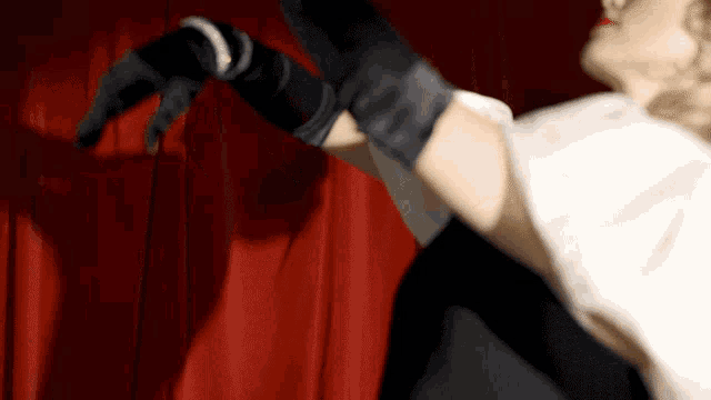a woman wearing black gloves and a white top is dancing in front of a red curtain