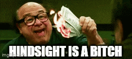 a man with glasses is holding a burning dollar bill and says hindsight is a bitch .