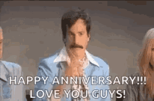 a man with a mustache is sitting in front of a group of people and saying `` happy anniversary ! `` .