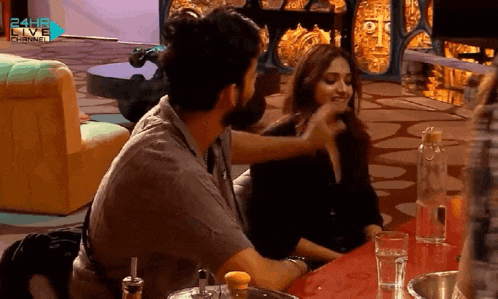 a man is touching a woman 's face while sitting at a table with a 24h6 live channel logo in the background