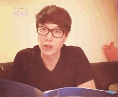 a man wearing glasses and a black shirt is sitting on a couch reading a book