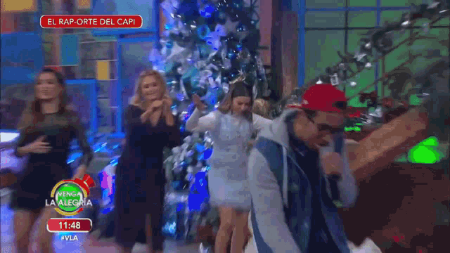 a woman in a white dress is dancing in front of a christmas tree while a man in a red hat looks on