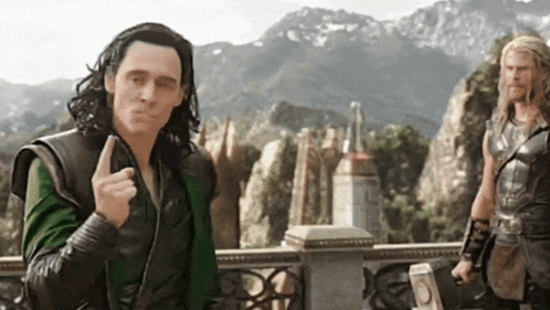 a man with long hair is giving a thumbs up while standing in front of a mountain .