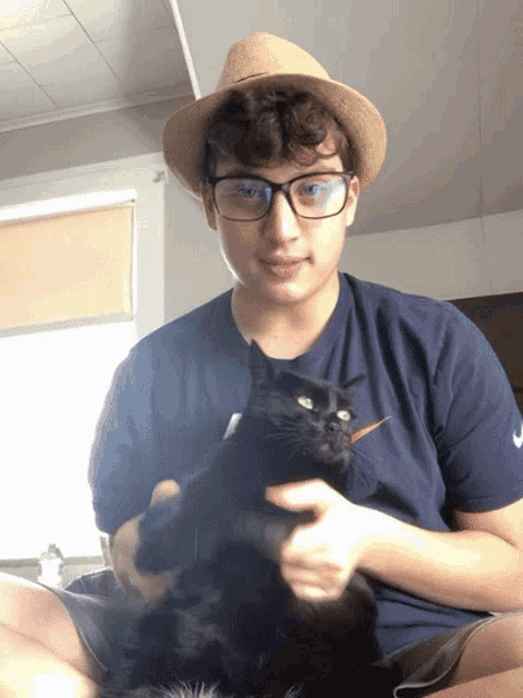 a young man wearing glasses and a hat holds a black cat