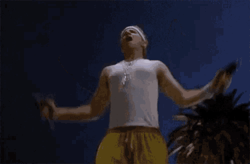 a man in a white tank top and yellow shorts is standing with his arms outstretched in front of a palm tree .