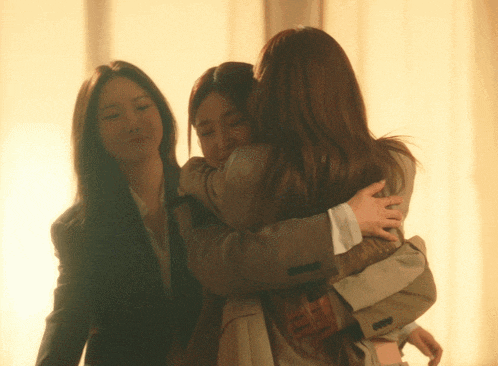 three women are hugging each other in front of a window and one of them is wearing a suit