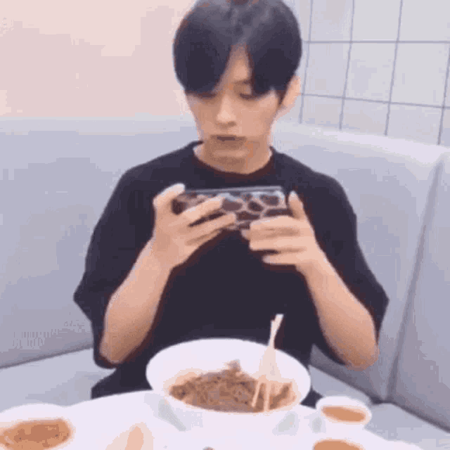 a young man is sitting at a table eating noodles and looking at his phone .