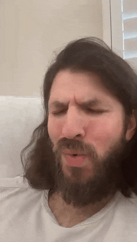 a man with long hair and a beard is sitting on a couch and making a funny face .