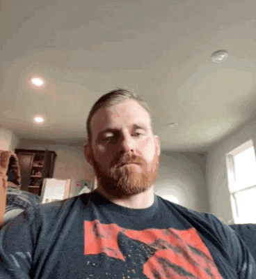 a man with a beard is sitting on a couch in a living room wearing a blue shirt .