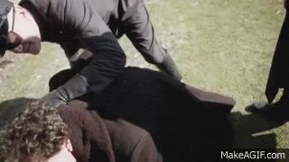 a man is laying on top of another man in a field while a group of people are standing around .