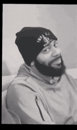 a man with a beard wearing a beanie and a hoodie is sitting on a couch .
