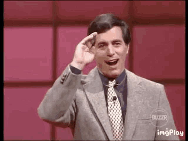 a man in a suit and tie is standing in front of a red wall and making a funny face .