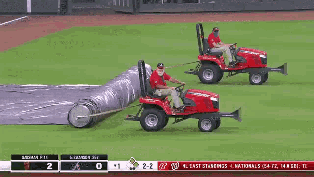 two men on tractors on a baseball field with a scoreboard behind them