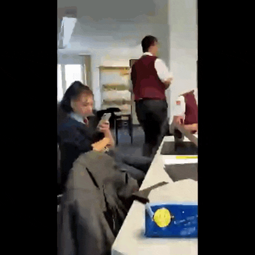 a man in a red vest is standing in a room with people sitting around a table