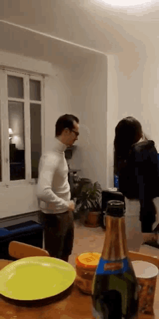 a man in a white turtleneck is standing in a living room next to a bottle of champagne .