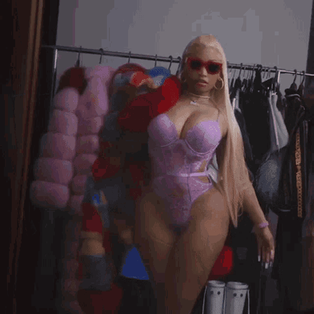 a woman wearing sunglasses and a pink bodysuit stands in front of a clothes rack